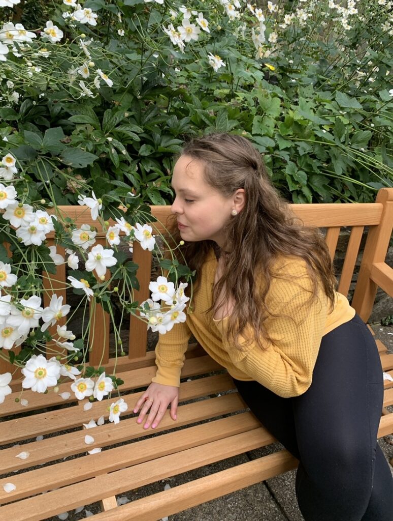 A picture of a girl in a yellow sweater and black leggings smelling white flowers while sitting on a brown bench. Her eyes are closed. 